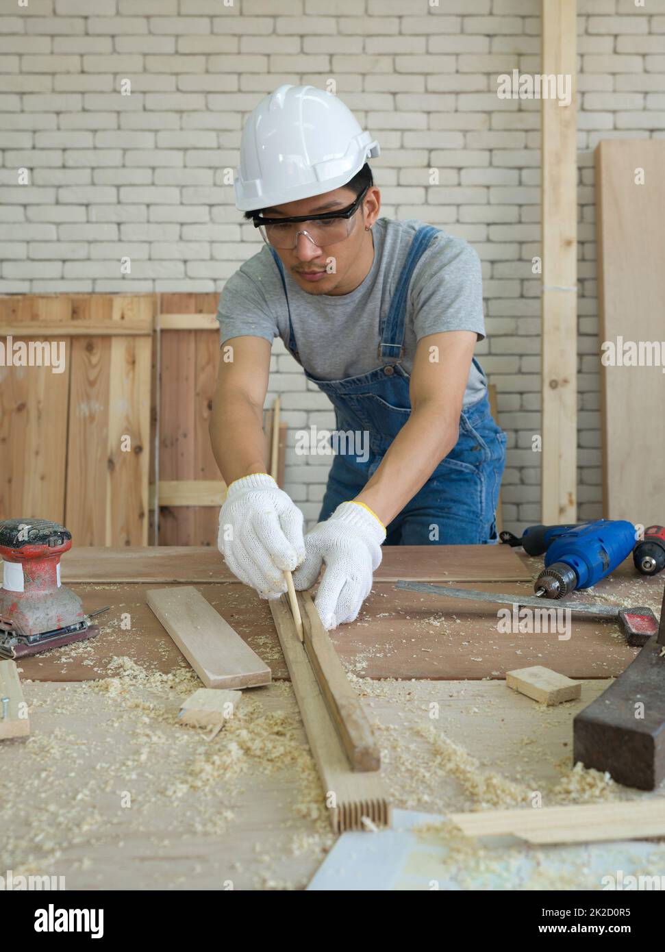 Charpentier asiatique avec des lunettes résistant à la poussière traçant une ligne sur un morceau de bois, se préparant à couper en sections. Ambiance de travail matinale dans la salle d'atelier. Banque D'Images