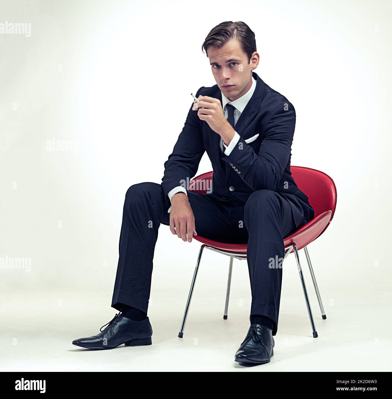 Suave fumeur. Un portrait en studio d'un beau jeune homme en costume à fines rayures assis sur une chaise. Banque D'Images