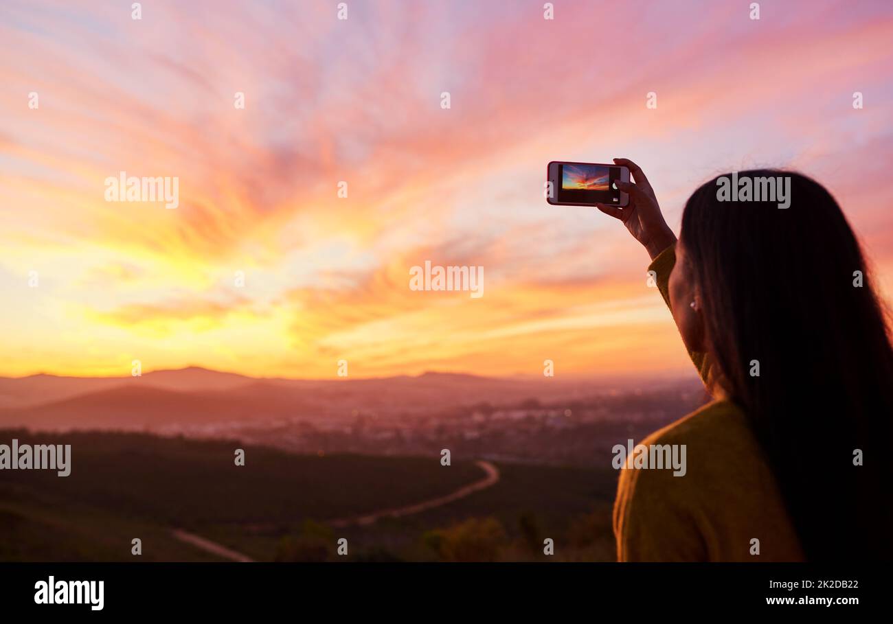 Je veux me souvenir de cela pour toujours. Prise de vue en vue arrière d'une femme prenant une photo d'un coucher de soleil avec son smartphone. Banque D'Images