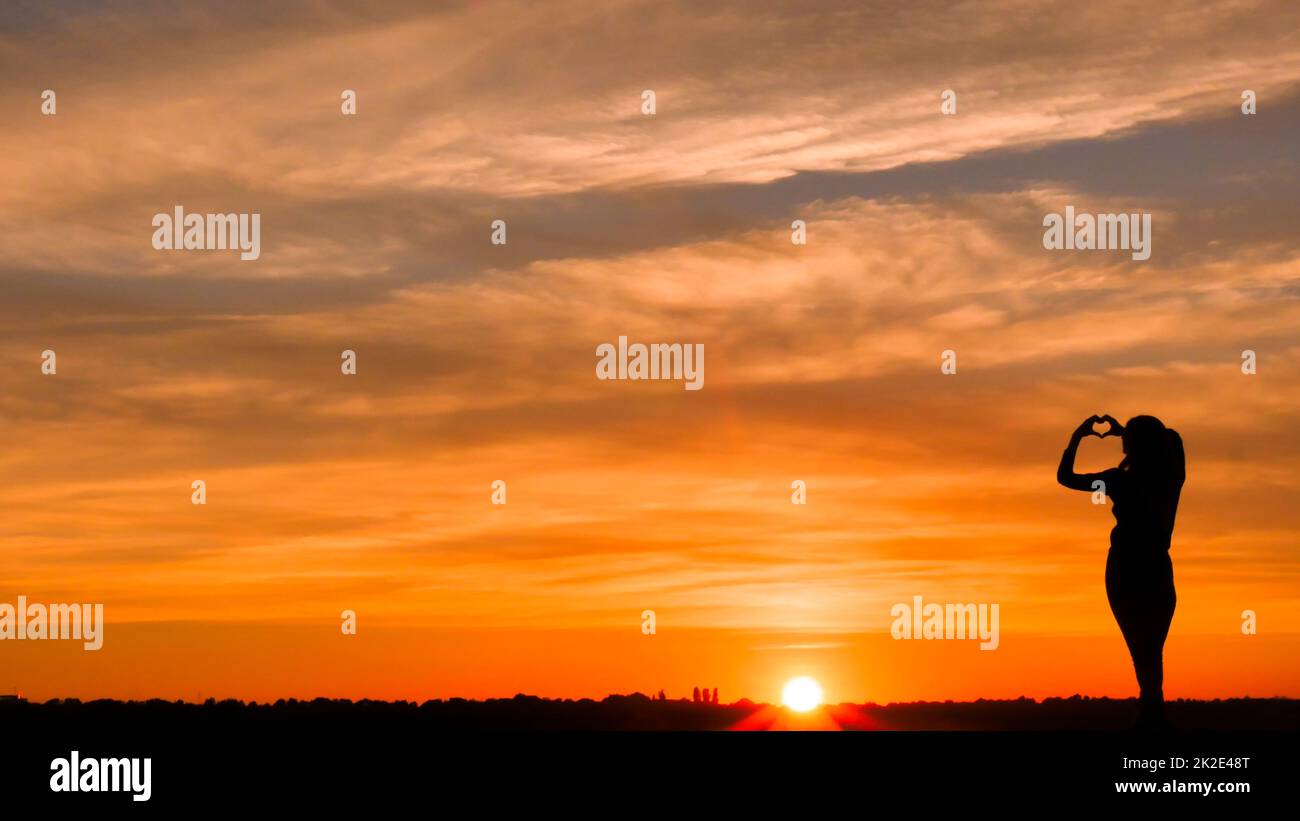 Femme en amour faisant un symbole de coeur à partir des doigts dans un ciel de feu arrière-plan. Banque D'Images