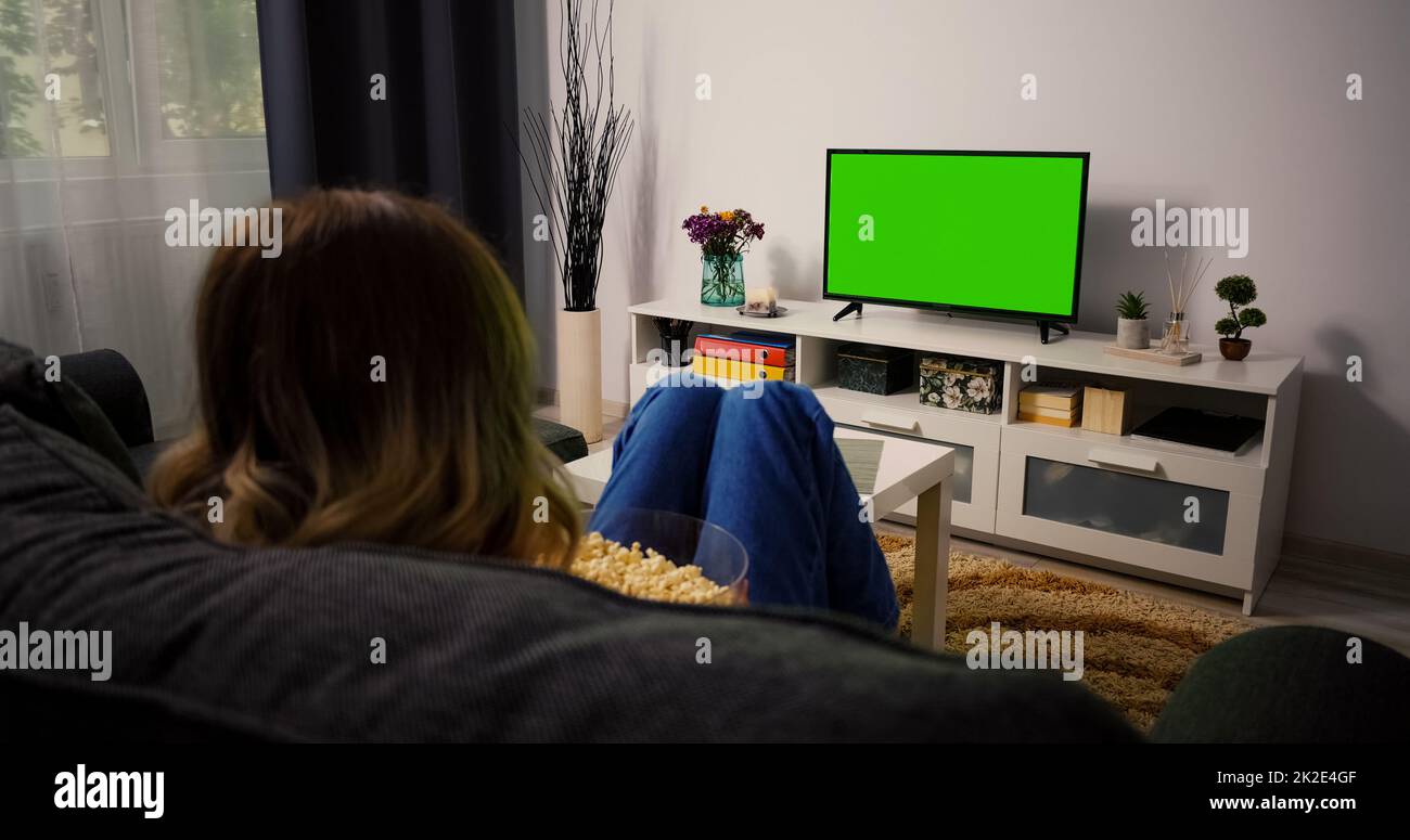 Femme assise sur un canapé à la maison regardant l'écran vert Chroma Key, se détendre. Une fille dans une chambre confortable regardant un match sportif, des nouvelles, un spectacle de télévision sitcom ou un film sur écran vert mangeant du pop-corn. Banque D'Images