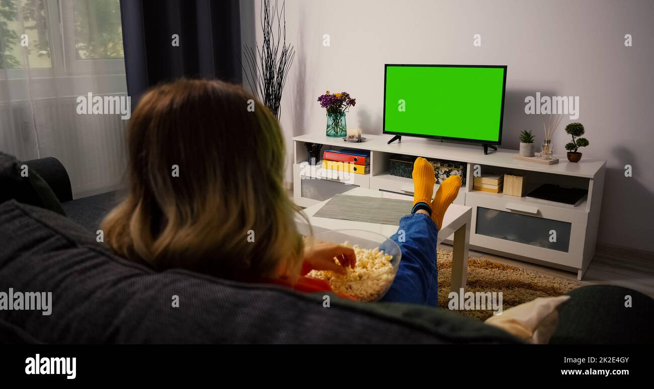 Femme regardant un téléviseur à écran couleur vert sur écran clé, se détendre. Une fille dans une chambre confortable regardant un match sportif, des nouvelles, un spectacle de télévision sitcom ou un film sur écran vert mangeant du pop-corn Banque D'Images