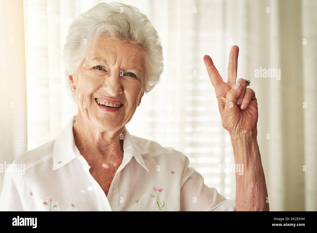 V pour la vitalité. Portrait d'une femme âgée heureuse montrant un geste de paix à la maison. Banque D'Images