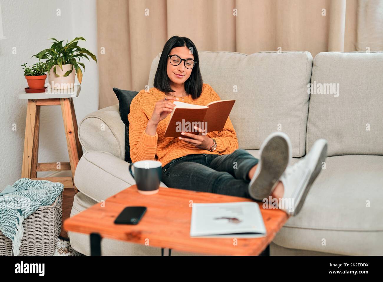 Les avantages de l'étude à la maison. Photo d'un jeune lisant un livre tout en travaillant sur le canapé à la maison. Banque D'Images