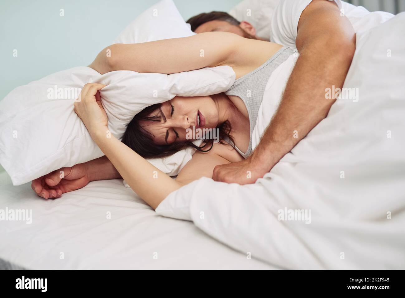 Je veux juste dormir en paix. Photo d'une jeune femme qui a l'air mal à l'aise et qui met un oreiller sur sa tête pour essayer de dormir pendant que son partenaire ronfle dans le lit. Banque D'Images