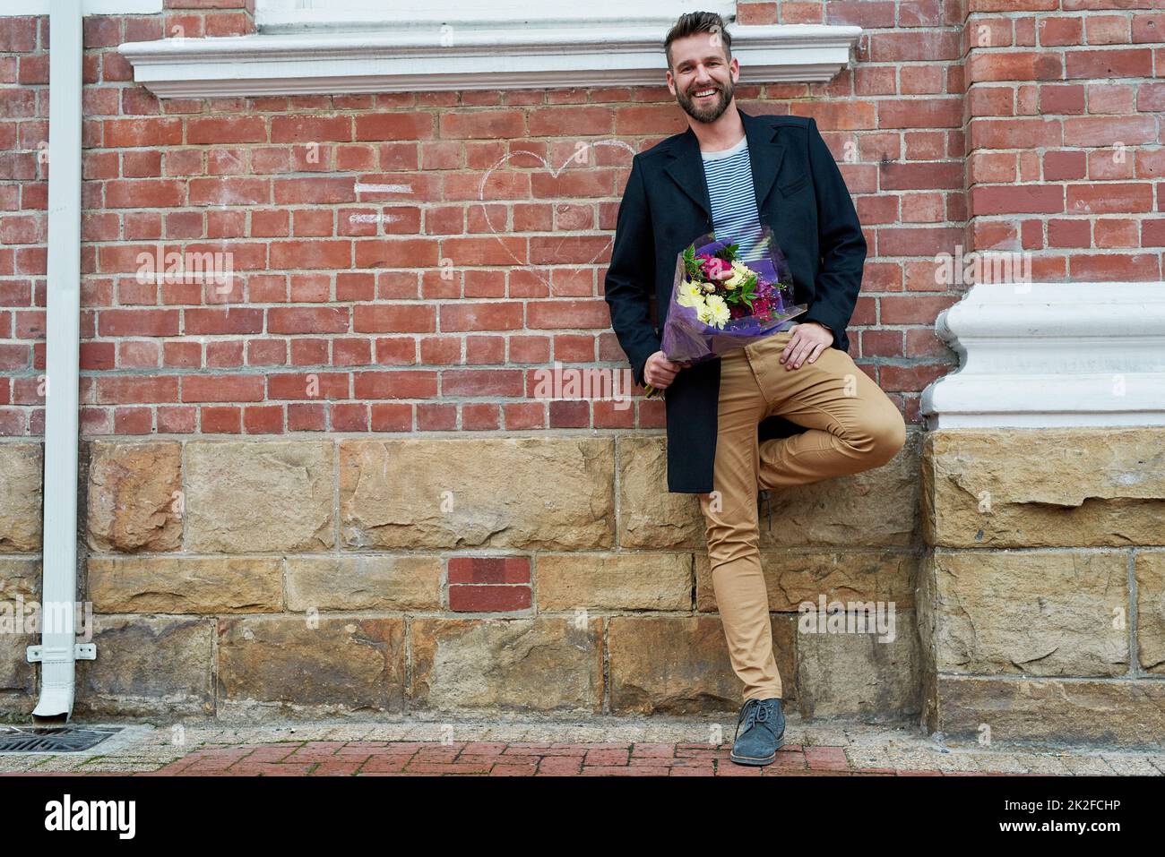 C'est pour l'amour de ma vie. Portrait d'un beau jeune homme debout contre un mur de briques avec un bouquet de fleurs à la main. Banque D'Images