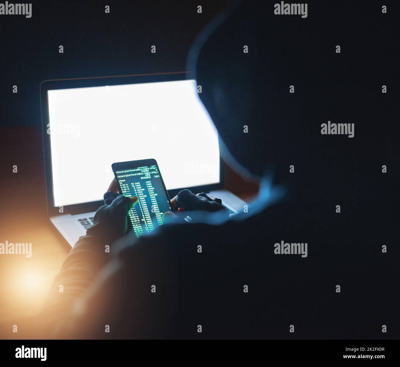 Accès à tous vos mots de passe. Photo d'un pirate méconnaissable à l'aide d'un téléphone portable et d'un ordinateur portable dans l'obscurité. Banque D'Images