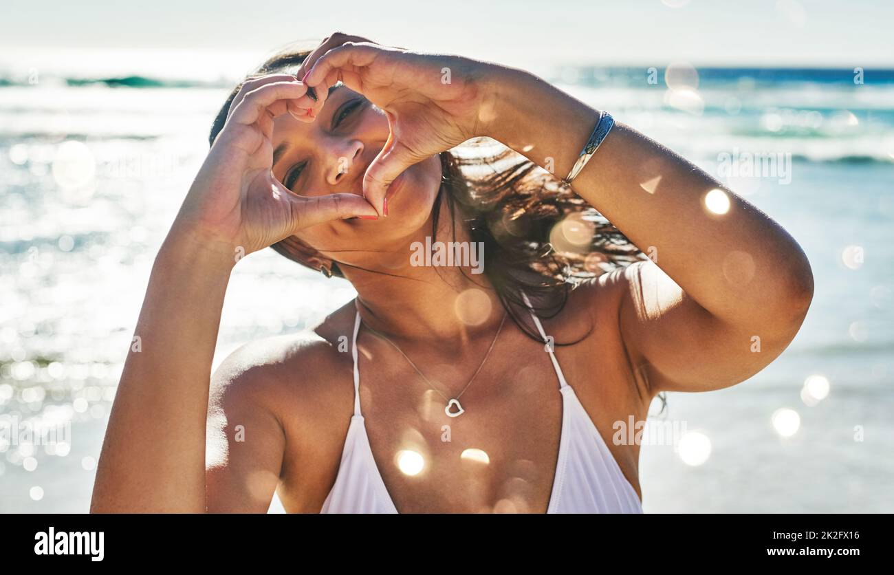 J'aime les gens qui aiment l'été. Portrait d'une belle jeune femme faisant un geste en forme de coeur avec ses mains à la plage. Banque D'Images