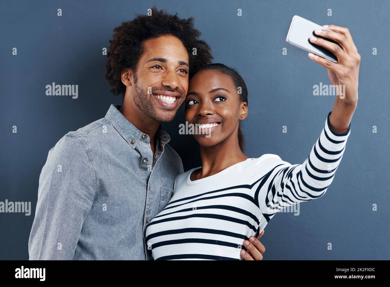 Nouvelle photo de profil. Photo d'un jeune couple heureux prenant une photo d'eux-mêmes avec un téléphone portable sur fond gris. Banque D'Images