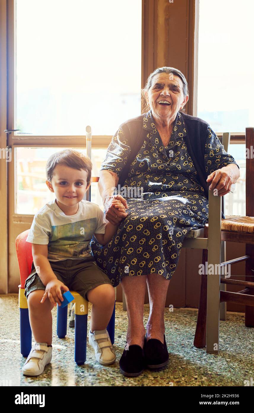 La jeunesse et les sages. Portrait d'un petit garçon joyeux assis à côté de sa grande grand-mère tout en regardant dans l'appareil photo à la maison. Banque D'Images