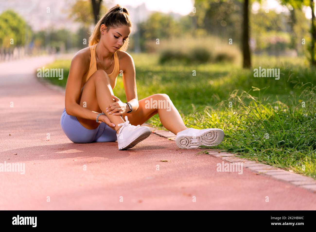 Portrait d'une femme de 20s sur une piste de course avec des hauts jaunes et des collants bleus tenant sa cheville dans la douleur. Banque D'Images