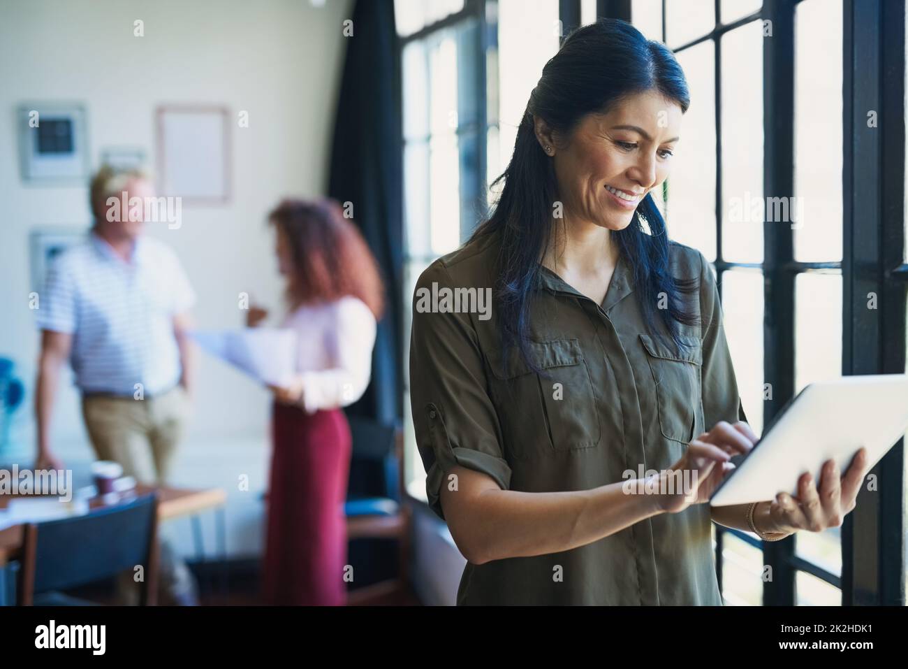 Gestion à l'ère de l'application. Photo d'une femme mûre utilisant une tablette numérique avec son équipe dans le fond d'un bureau moderne. Banque D'Images