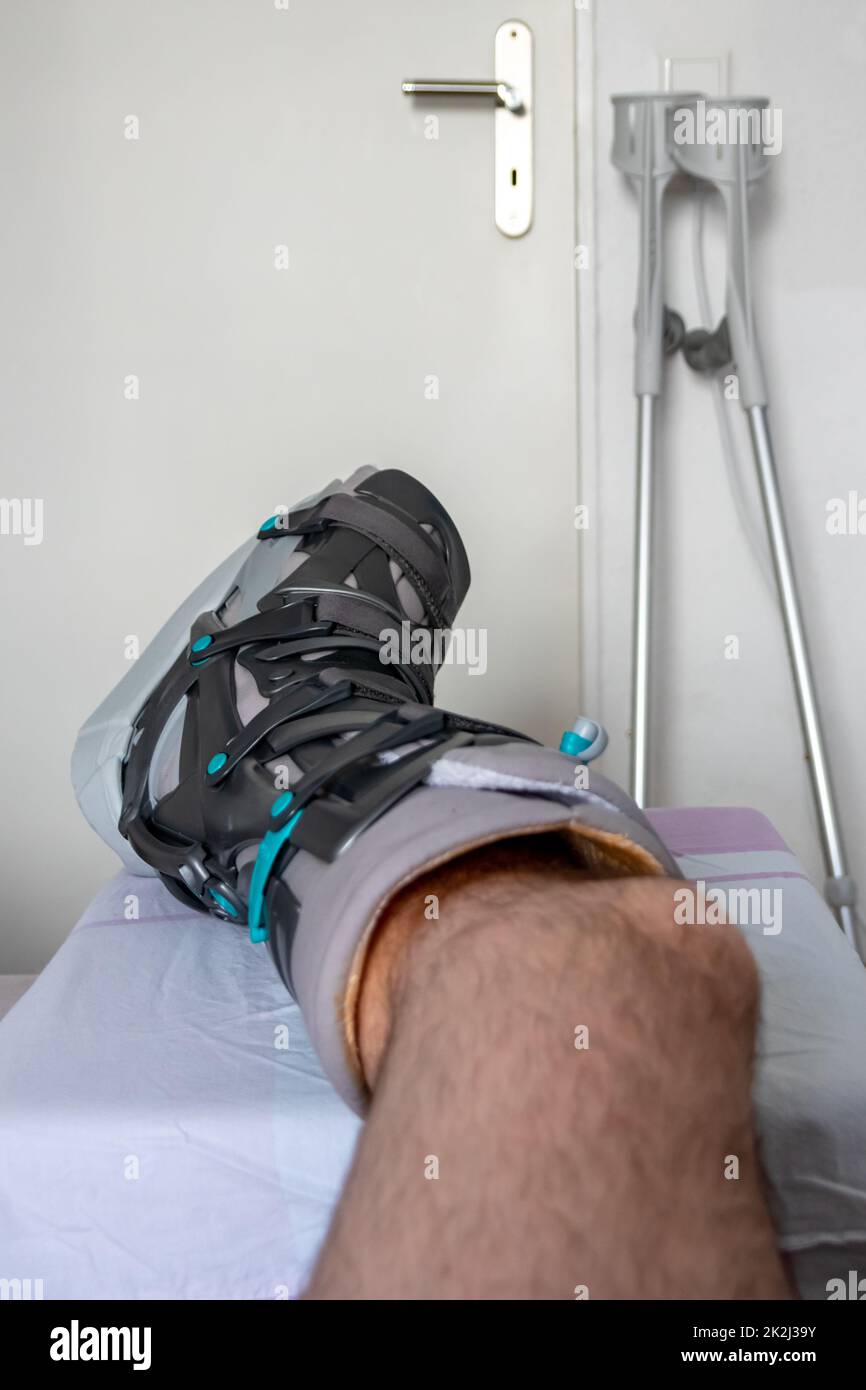 L'homme européen après l'opération de rupture de tendon d'Achille est de retour à la maison avec des chaussures spéciales de physiothérapie et des béquilles pour la récupération à la maison avec la médecine saine analgésique médicaments pilules contre la jambe blessant Banque D'Images