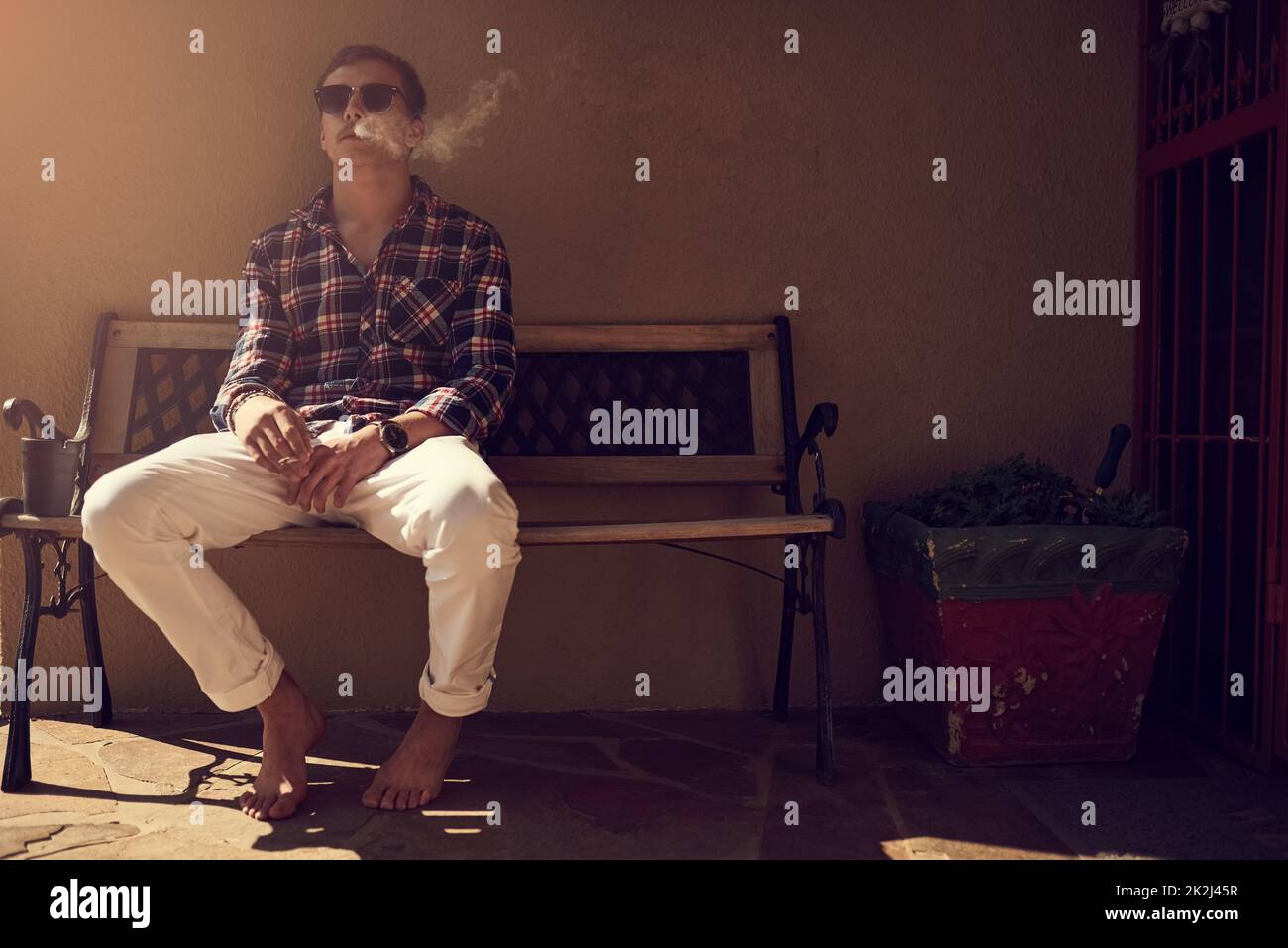 Prendre une bouffée dans la véranda. Photo d'un jeune homme assis sur un banc à l'extérieur et fumant une cigarette. Banque D'Images