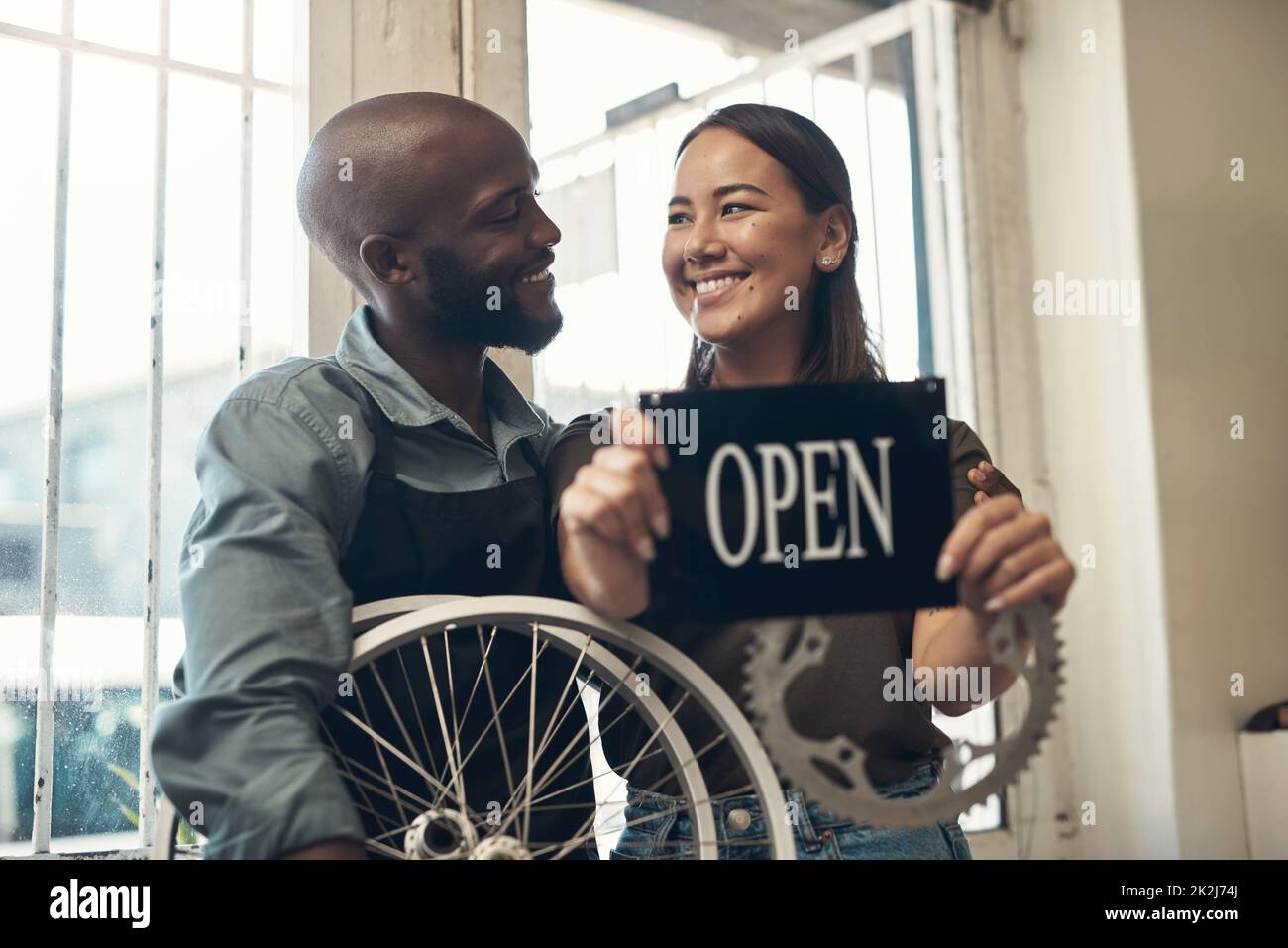 Je suis si fière de son entreprise. Photo de deux jeunes propriétaires d'entreprise se tenant ensemble dans leur magasin de vélos et tenant un panneau ouvert. Banque D'Images