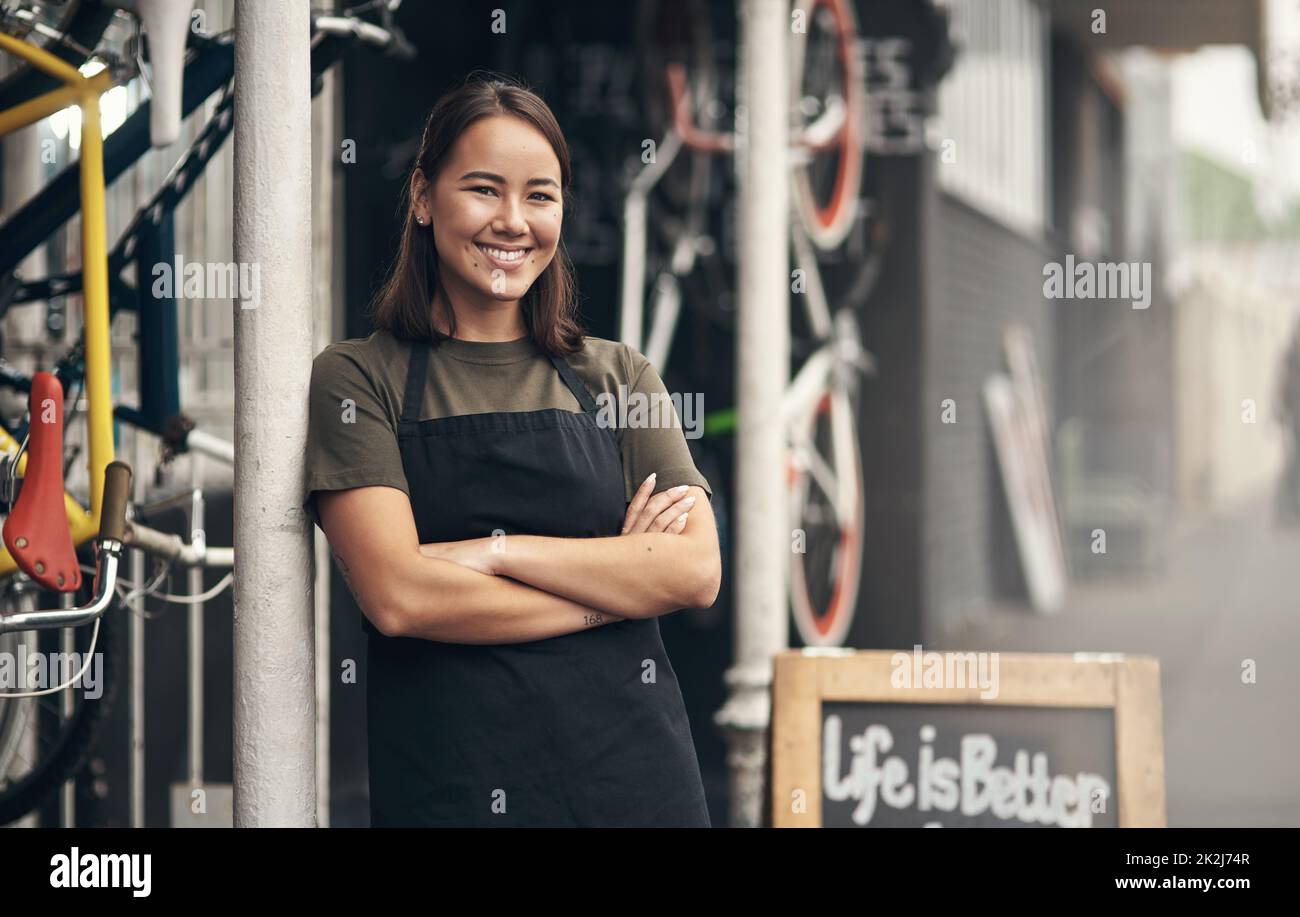 Demandez-moi quelque chose sur les vélos. Prise de vue d'une jeune femme attrayante debout à l'extérieur de son magasin de vélos avec ses bras pliés. Banque D'Images