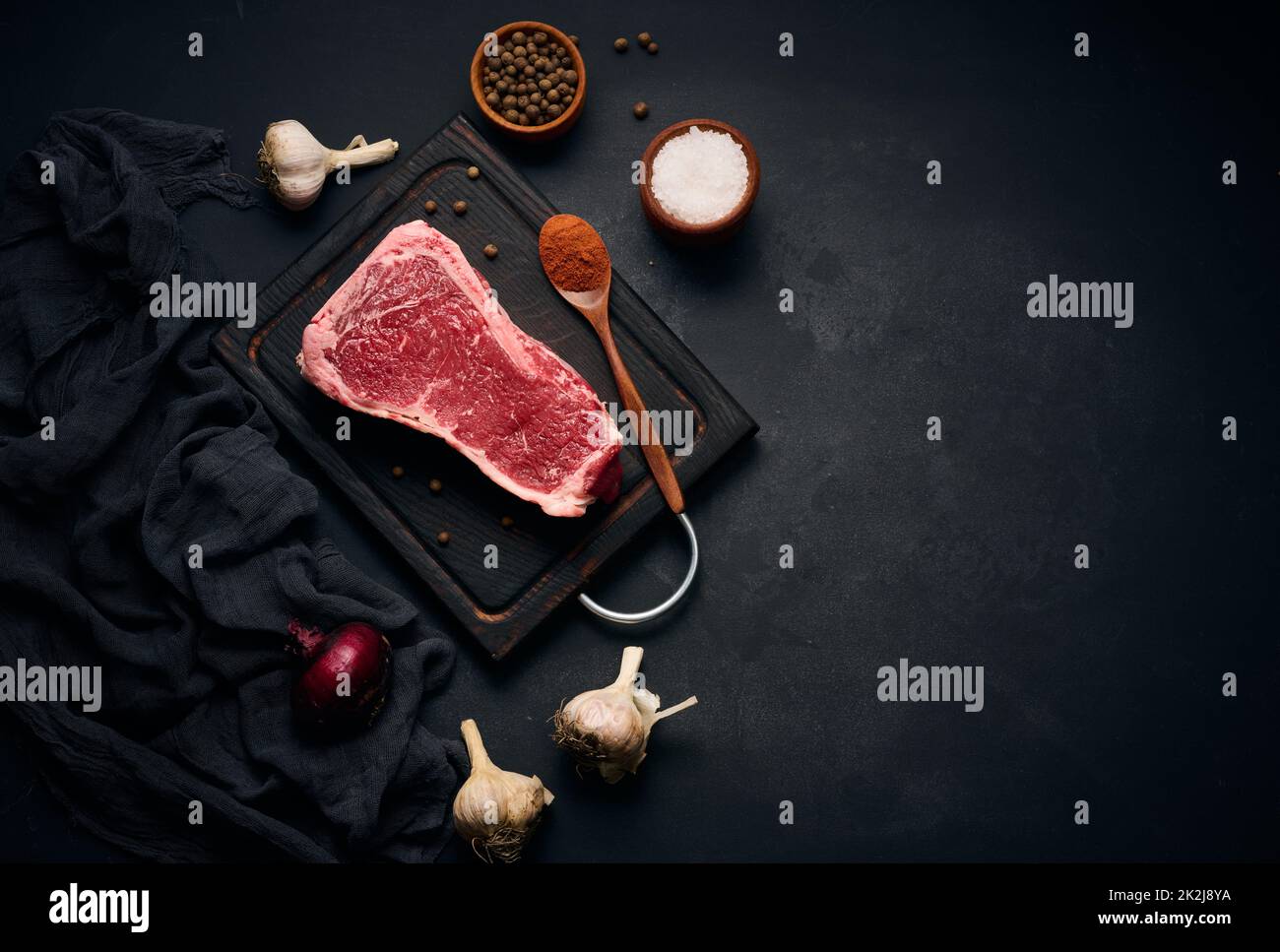 Morceau de viande de bœuf cru juteux sur l'os repose sur une planche à découper en bois, épices pour cuisiner sur fond noir. Filet de viande New York Banque D'Images