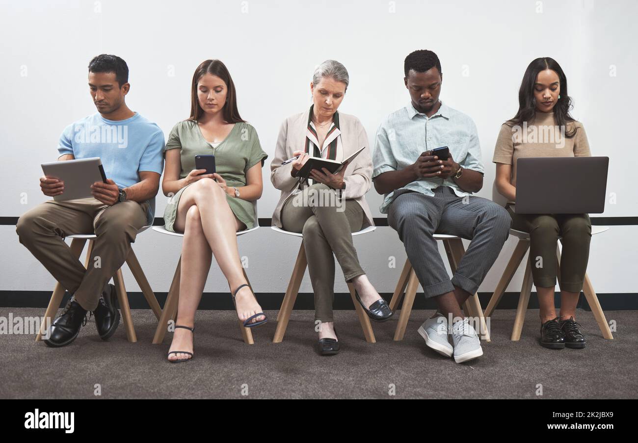 Rester concentré pendant qu'ils se préparent chacun à quelque chose de grand. Photo d'un groupe d'hommes d'affaires utilisant des appareils numériques tout en étant assis ensemble dans une ligne contre un mur blanc. Banque D'Images