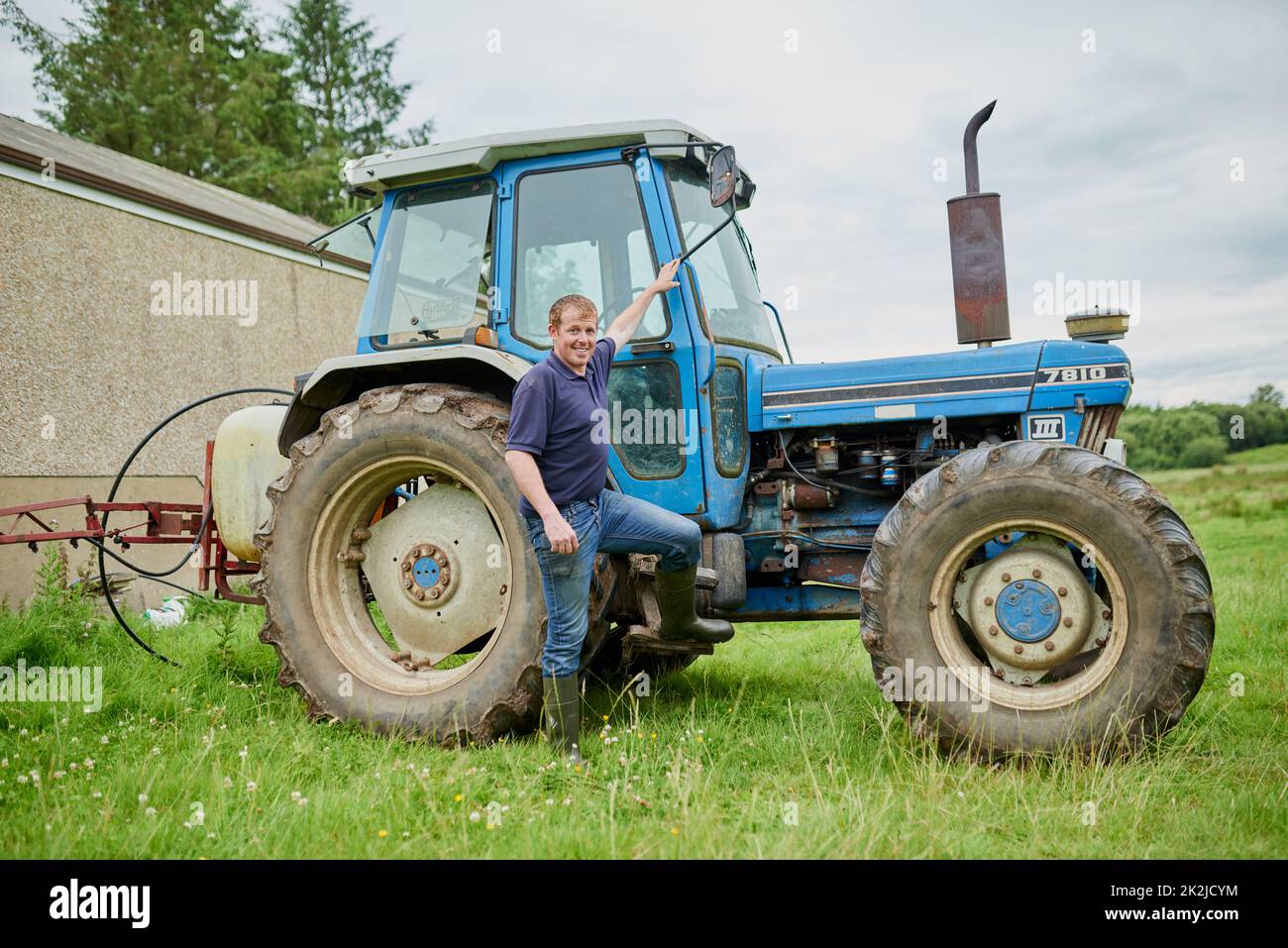 Venez jeter un œil dans mon bureau. Portrait d'un jeune agriculteur joyeux qui se pose à côté de son grand tracteur agricole à l'extérieur sur un champ. Banque D'Images
