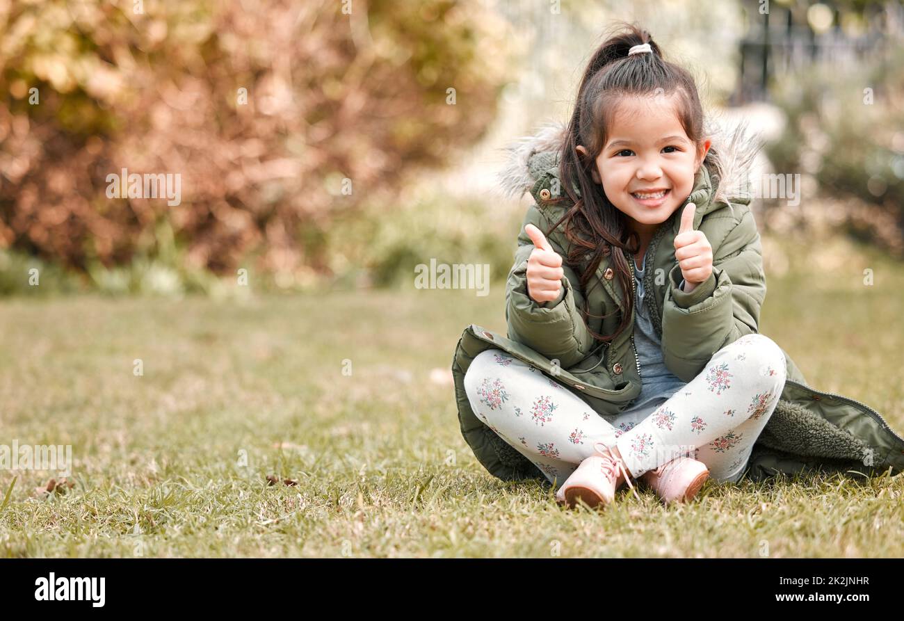 J'aime ça ici. Photo d'une adorable petite fille montrant les pouces vers le haut tout en étant assis à l'extérieur. Banque D'Images