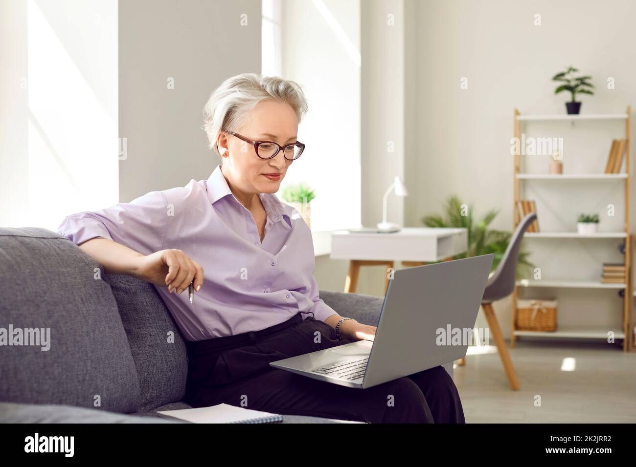 Femme âgée assise avec un ordinateur portable sur un canapé à la maison et ayant un appel vidéo ou regardant un séminaire en ligne Banque D'Images