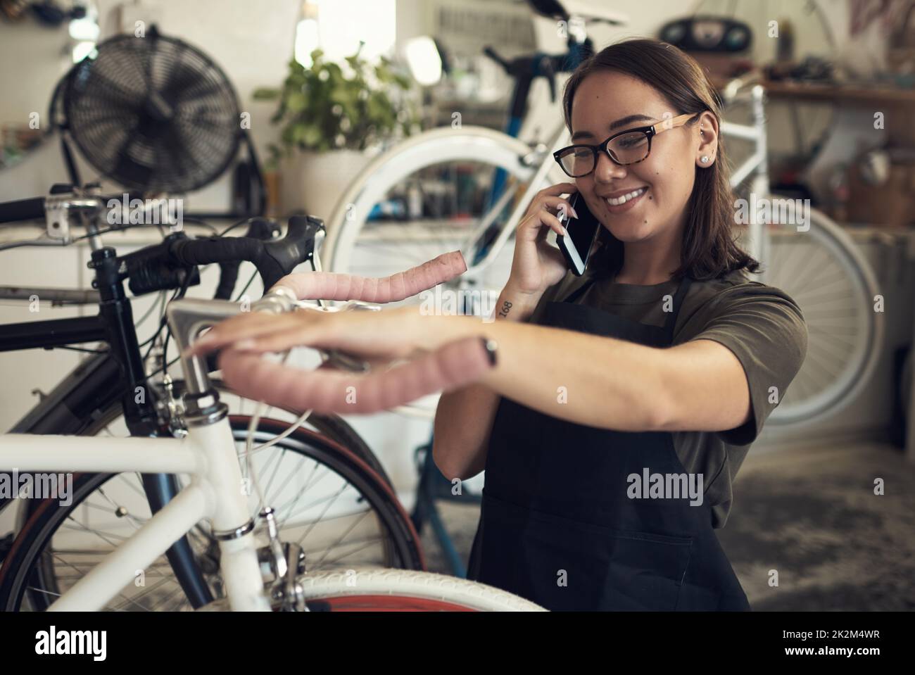 J'ai préparé votre vélo pour le ramassage. Photo d'une jeune femme attrayante debout seule dans son magasin et en train de réparer une roue de vélo tout en utilisant son téléphone portable. Banque D'Images