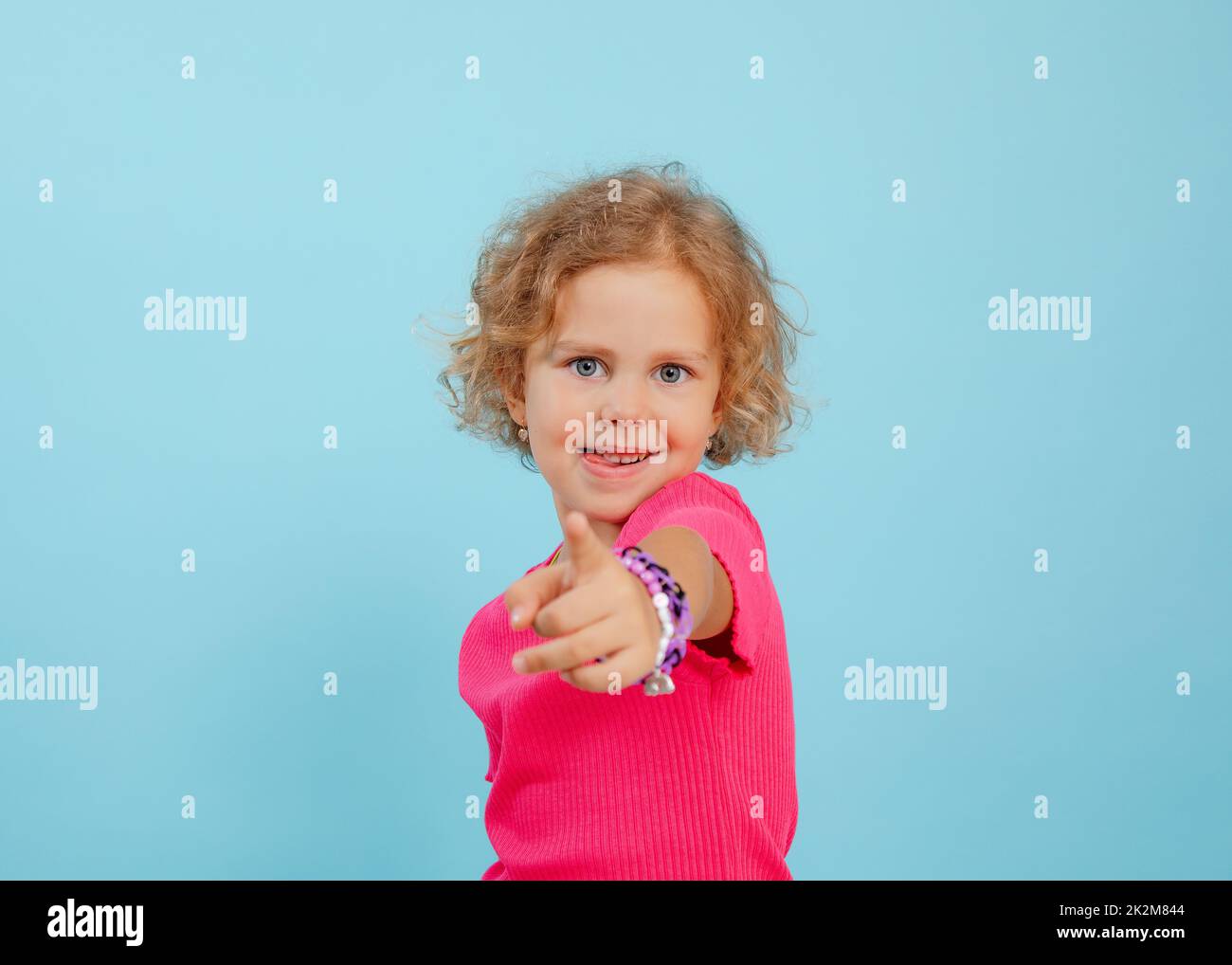 Portrait d'une petite fille aux yeux bleus avec des cheveux bouclés et justes, pointant vers l'appareil photo avec l'index sur fond bleu. Banque D'Images