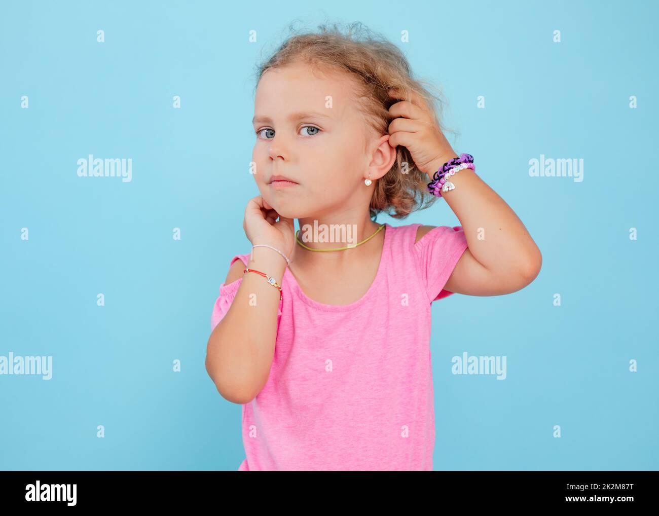 Portrait de chérubis bleu-eyed petite fille titivating touchant les cheveux courts bouclés justes, portant la combinaison rose, les bracelets. Banque D'Images