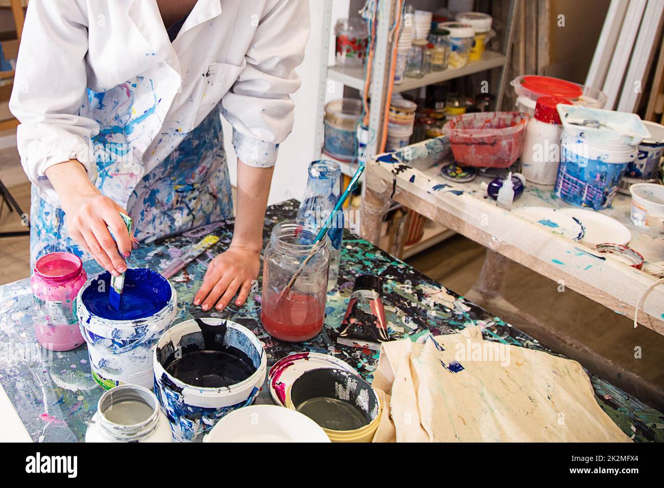 Photo rognée des mains d'une femme en robe recouverte de diverses taches remuant la peinture acrylique bleue dans le seau avec la brosse. Banque D'Images