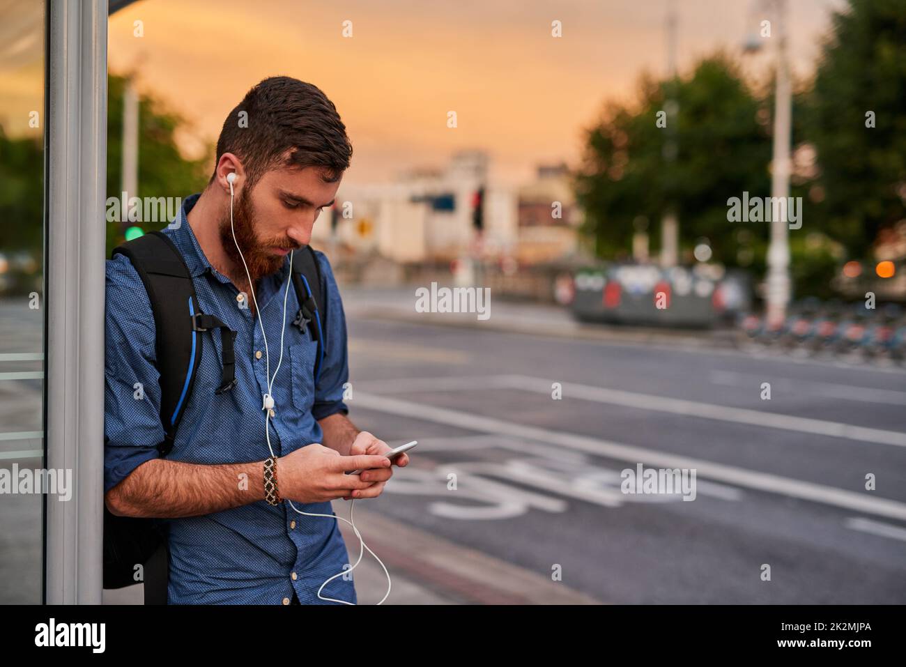 La musique fait toujours paraître l'attente plus courte. Photo rognée d'un beau jeune homme écoutant de la musique sur son téléphone portable en attendant un arrêt de bus. Banque D'Images