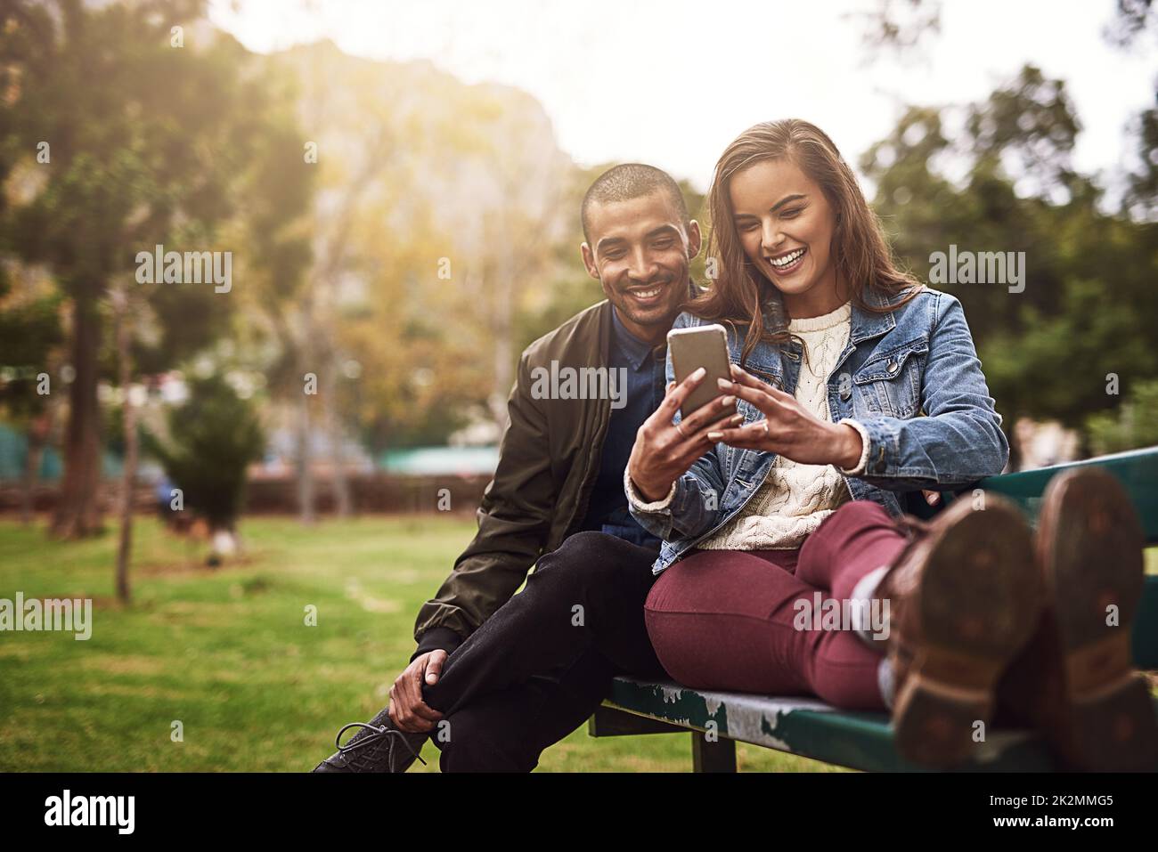 Que pensez-vous de cette photo ? Photo d'un jeune couple gai assis sur un banc tout en utilisant un téléphone ensemble à l'extérieur dans un parc. Banque D'Images