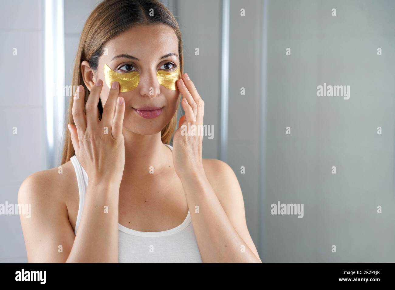 Femme de beauté appliquant l'anti-âge doré masque sous-oeil regardant elle-même dans le miroir dans la salle de bains. Soins de la peau une fille touche des timbres de tissu masque sous les yeux pour réduire les sacs pour les yeux. Banque D'Images