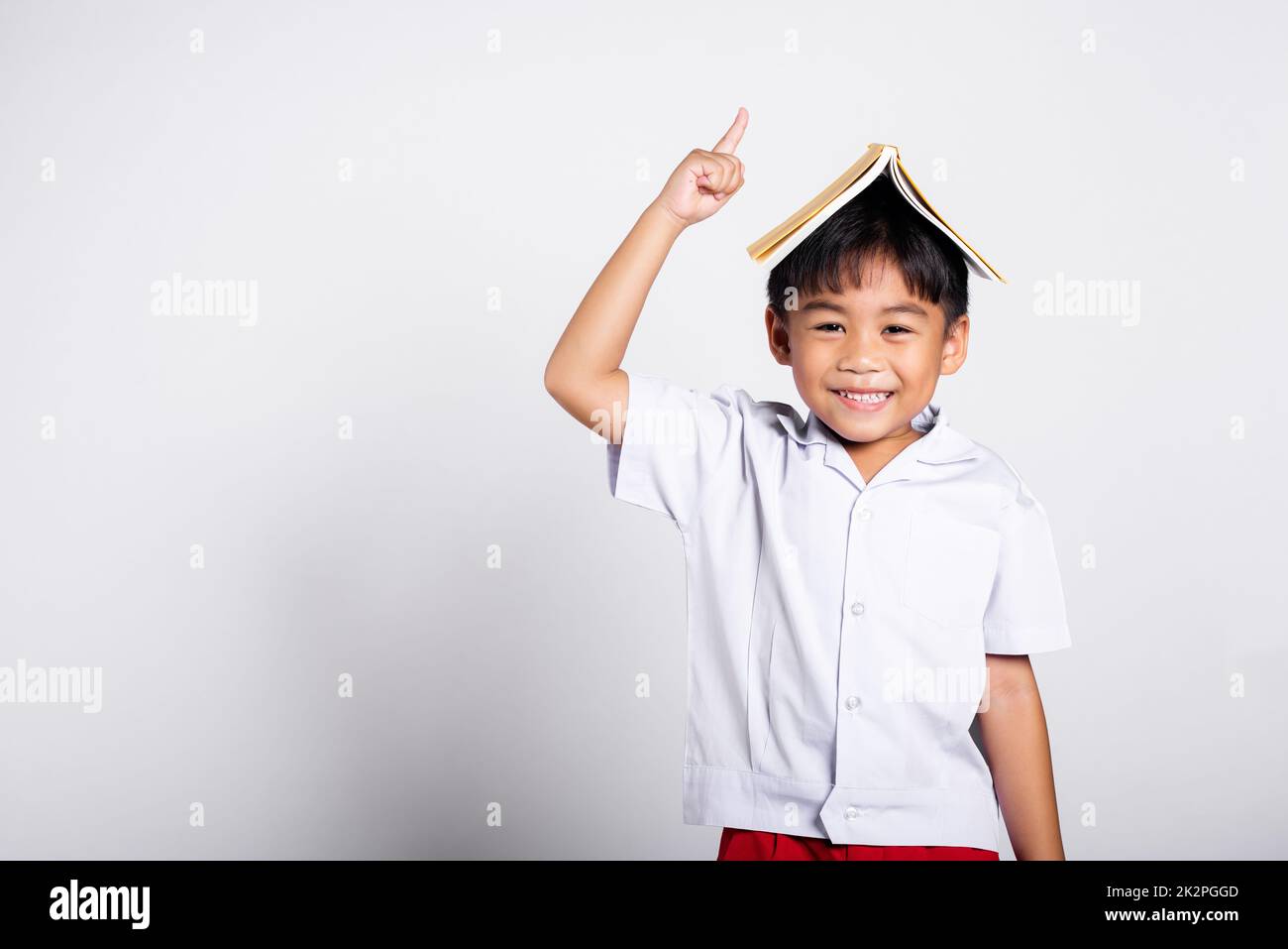 Asiatique adorable tout-petit souriant heureux portant étudiant thaïlandais uniforme pantalon rouge stand tenant le livre sur la tête comme le toit Banque D'Images