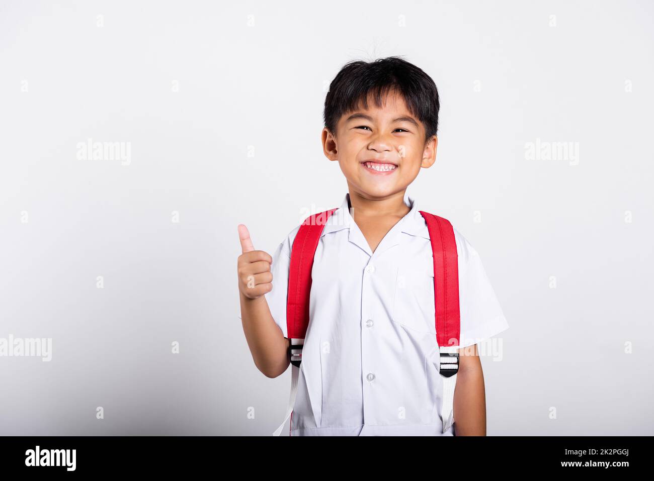 Asiatique adorable tout-petit souriant heureux portant étudiant thaïlandais uniforme pantalon rouge montrer pouce vers le haut de doigt Banque D'Images