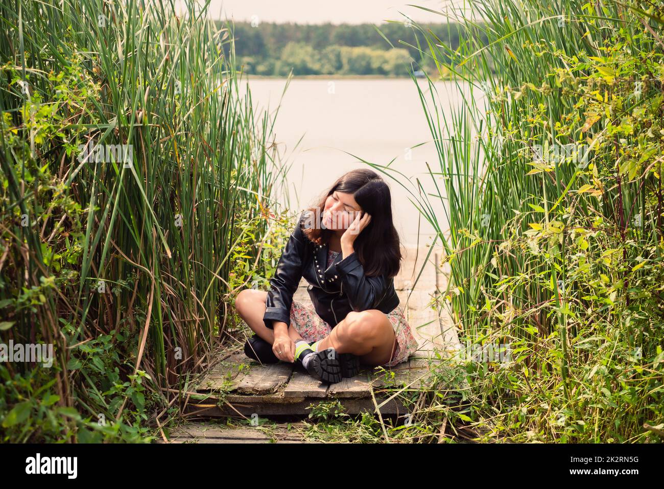 une fille brune dans une veste noire est assise sur un pont en bois sur la rive d'un étang, en la soutenant la tête vers le haut avec sa main Banque D'Images