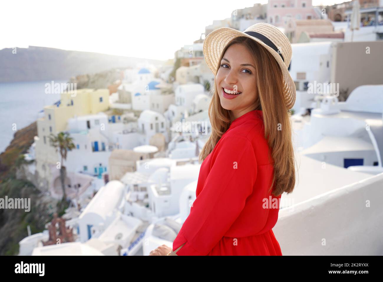 Bonne fille touristique appréciant le voyage en Grèce. Belle femme souriante et regardant la visite de caméra sur le coucher du soleil village d'Oia sur l'île de Santorini, Grèce, Europe. Banque D'Images