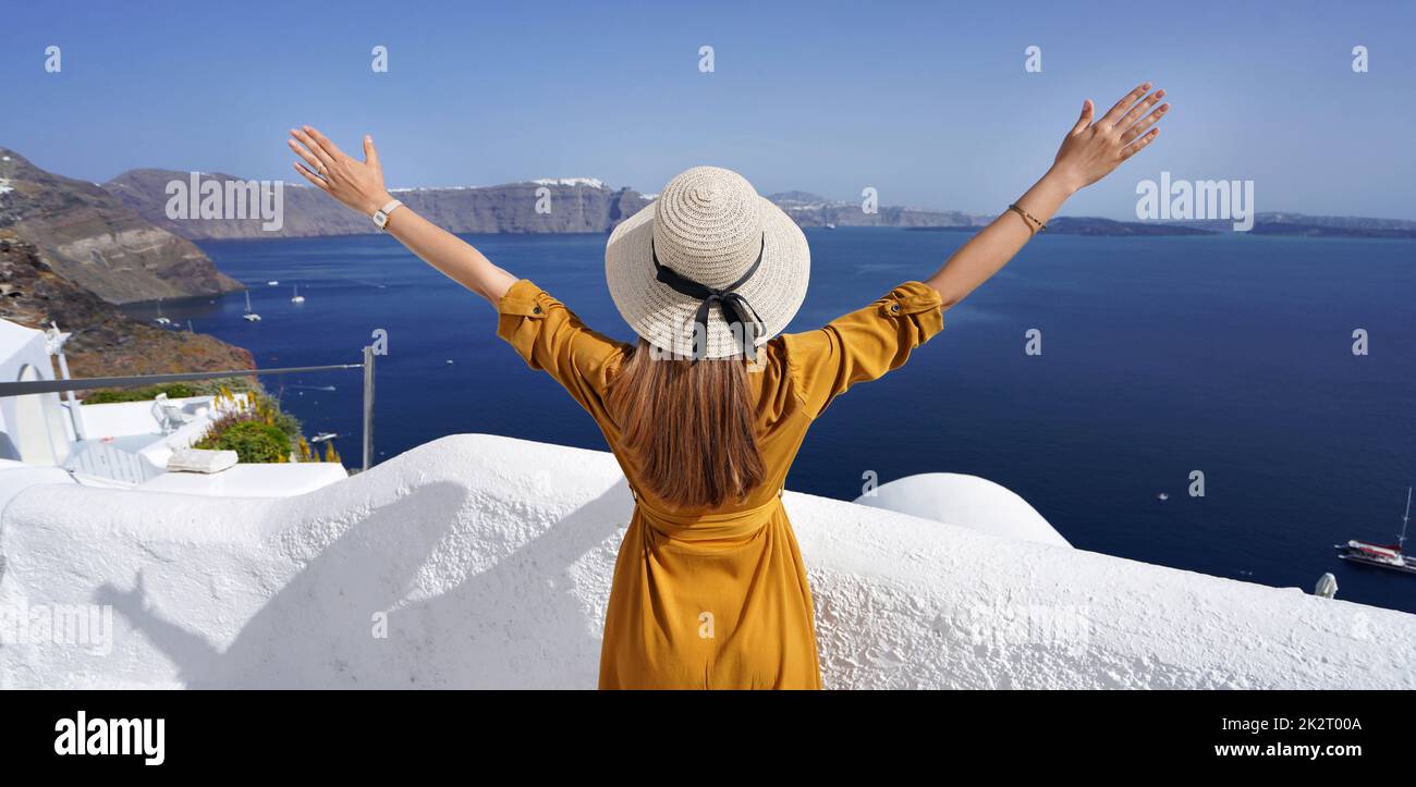 Europe Voyage femme avec les bras vers le haut regardant Santorini Caldera en Grèce. Une fille touristique insouciante dans une destination européenne portant une robe et un chapeau de mode. Banque D'Images