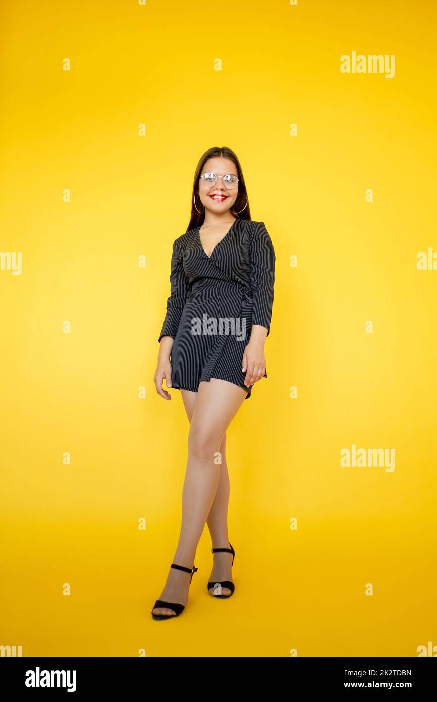 Style de vie de femme. Indépendance féminine. Confiance réussie. Gai élégant femme en verres mini robe souriante debout isolée sur jaune copie s. Banque D'Images