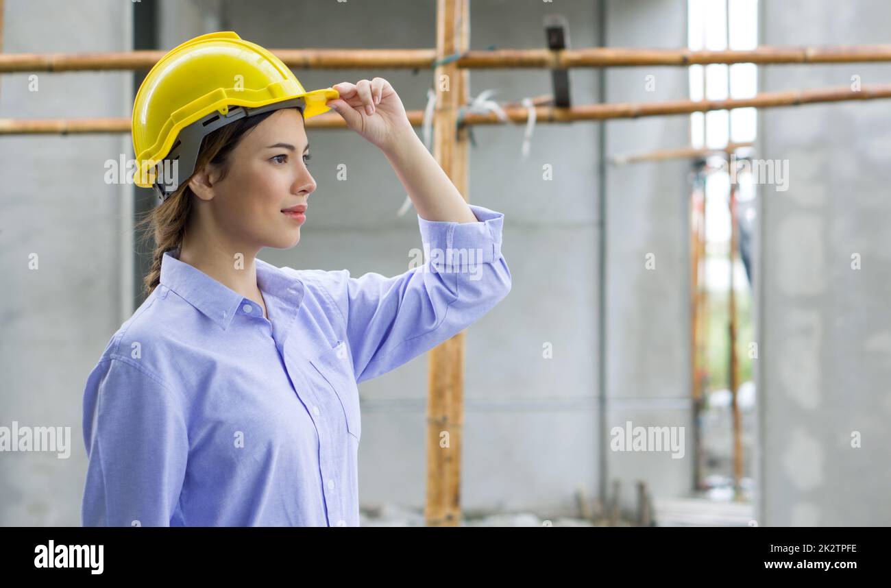 Jeune belle femme brun cheveux tenant casque de construction tout en regardant l'avancement des projets immobiliers. Le site de construction a des échafaudages en bois en arrière-plan. Banque D'Images