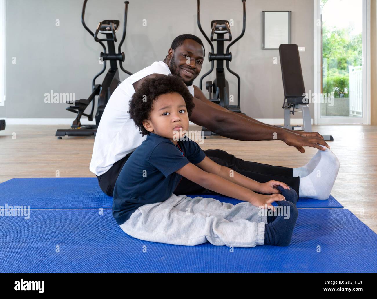 Jeune garçon de cheveux noir bouclés s'étirant avec son père après avoir terminé la routine d'exercice sur le tapis de yoga. Des appareils de cardio-training sont à l'arrière-plan à la salle de gym. Banque D'Images