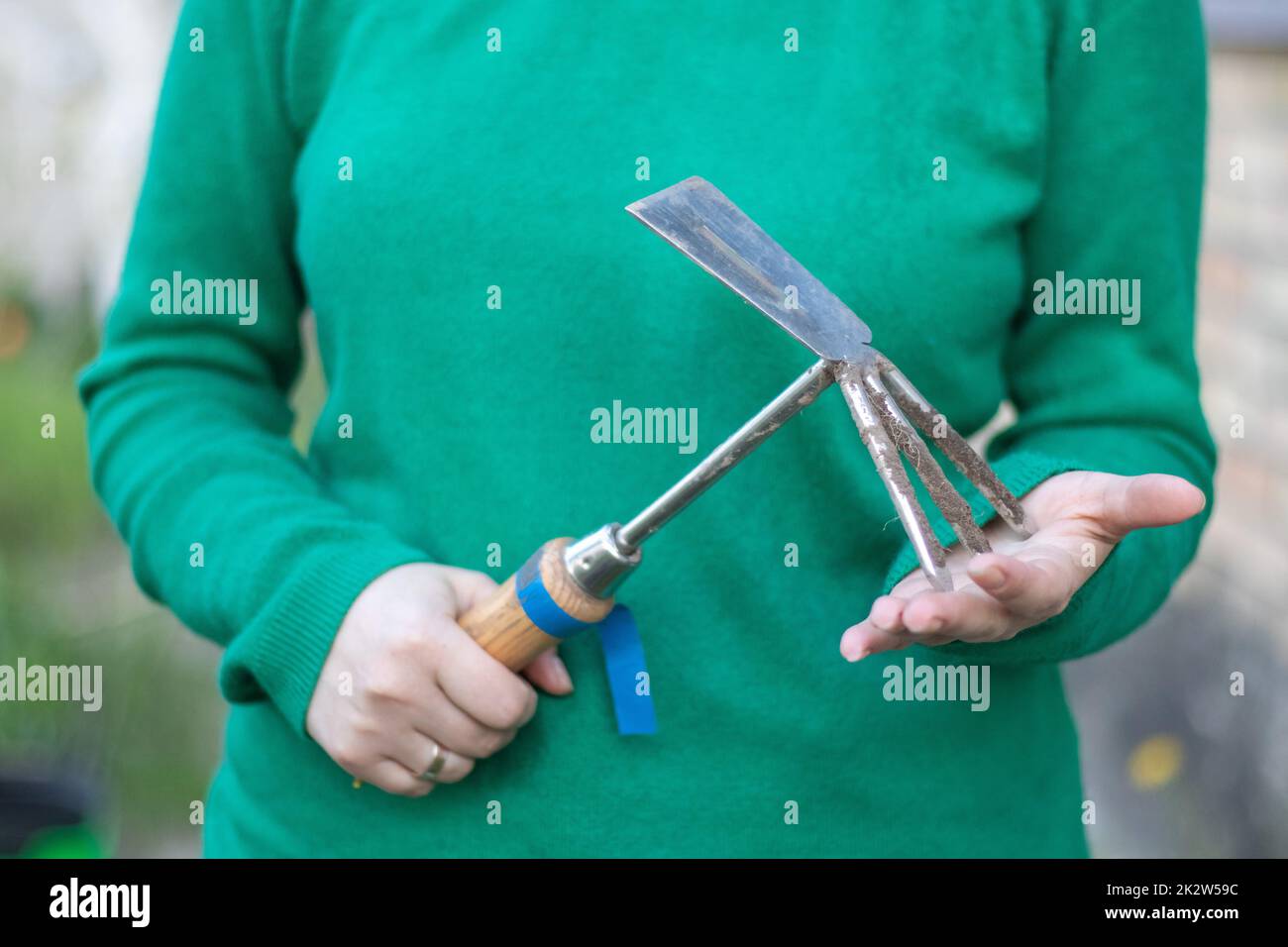 Une femme caucasienne jardinière dans le jardin montre un râteau. Le concept de jardinage et d'été durable. Main de femme avec un outil de jardin. Peigne à hache pour le jardinage dans les mains des femmes. Placer pour le texte. Banque D'Images