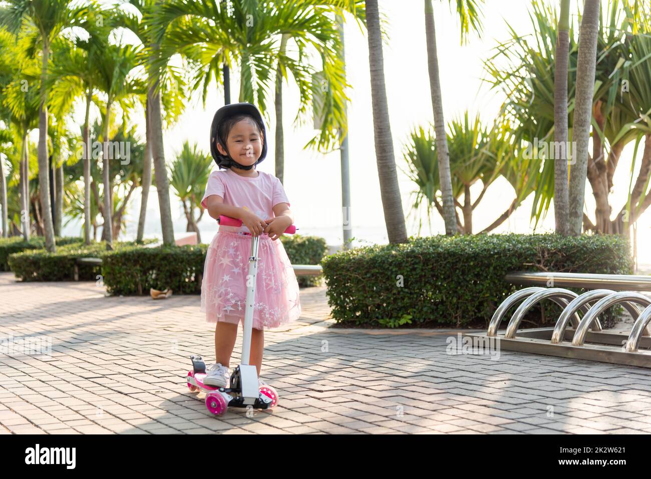 Joyeux asiatique petit enfant fille porter un casque sûr jouant de la planche à pied rose sur la route dans le parc à l'extérieur le jour d'été Banque D'Images