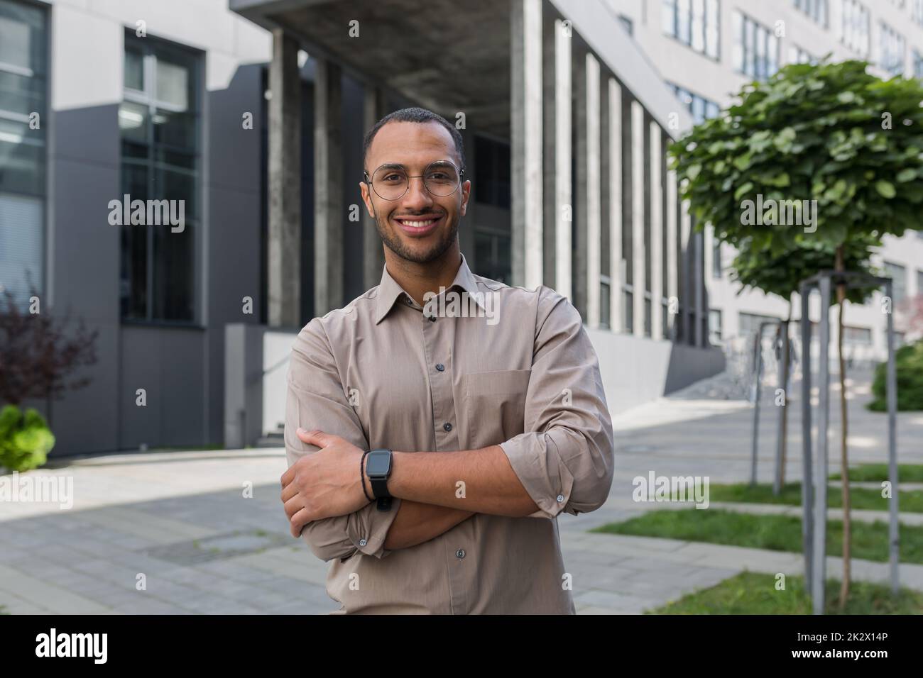 Jeune homme d'affaires réussi souriant et regardant la caméra avec des bras croisés, homme à l'extérieur du bâtiment de bureau portant une chemise et des lunettes, startup investisseur. Banque D'Images