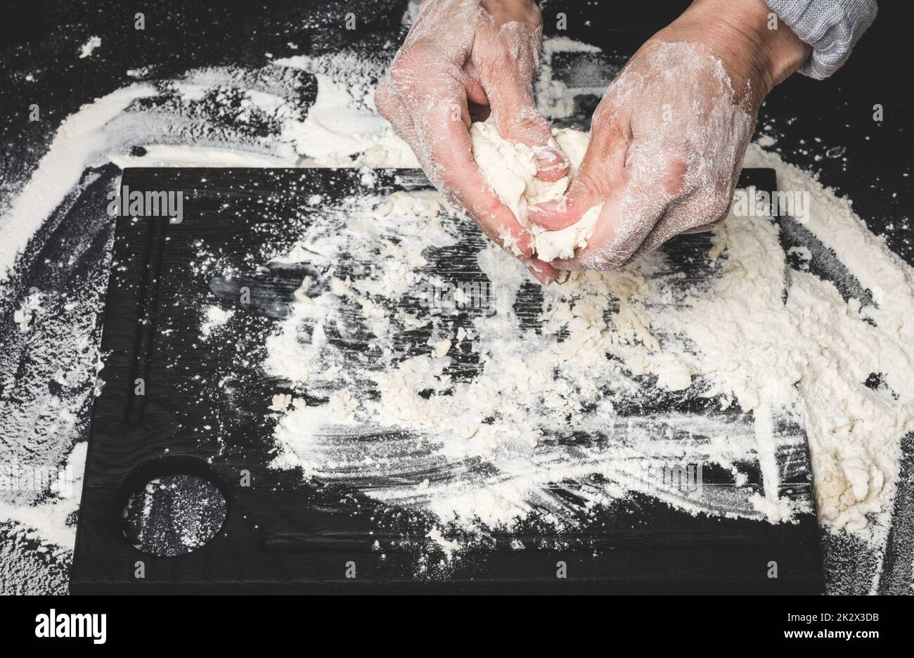 deux mains de femmes pétrient la pâte de farine de blé blanc sur une table noire, vue de dessus Banque D'Images