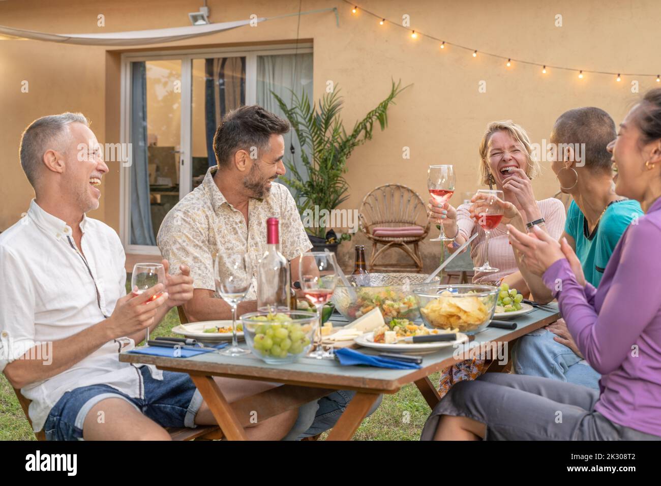 Amis multiethniques mangeant et buvant souriant et riant dans le patio. Personnes d'âge moyen et gaies autour de la table. Banque D'Images