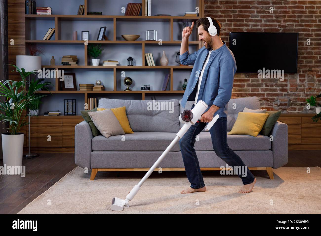 Homme joyeux, l'homme nettoie l'appartement. Passer l'aspirateur, porter un casque avec danser et chanter dans un smartphone au lieu d'un microphone. Maison Banque D'Images