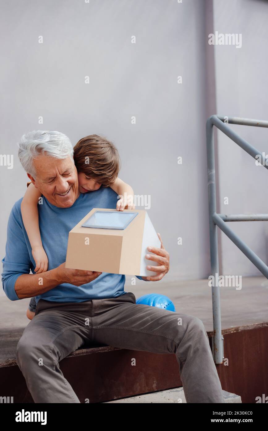 Homme senior souriant tenant un modèle de maison solaire par petit-fils devant le mur Banque D'Images