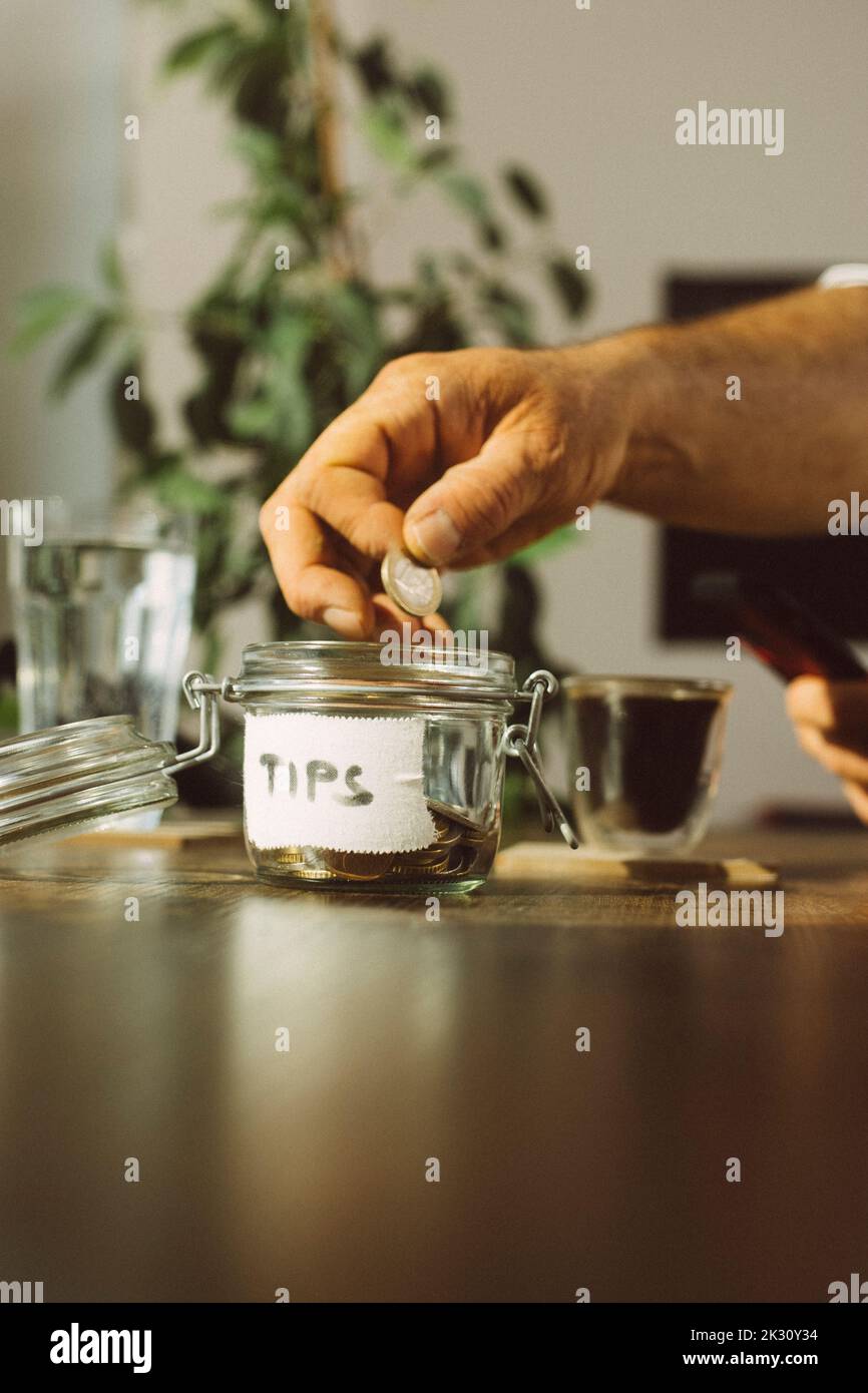 Main d'homme d'affaires mettant la pièce dans le pot de pourboire au café Banque D'Images