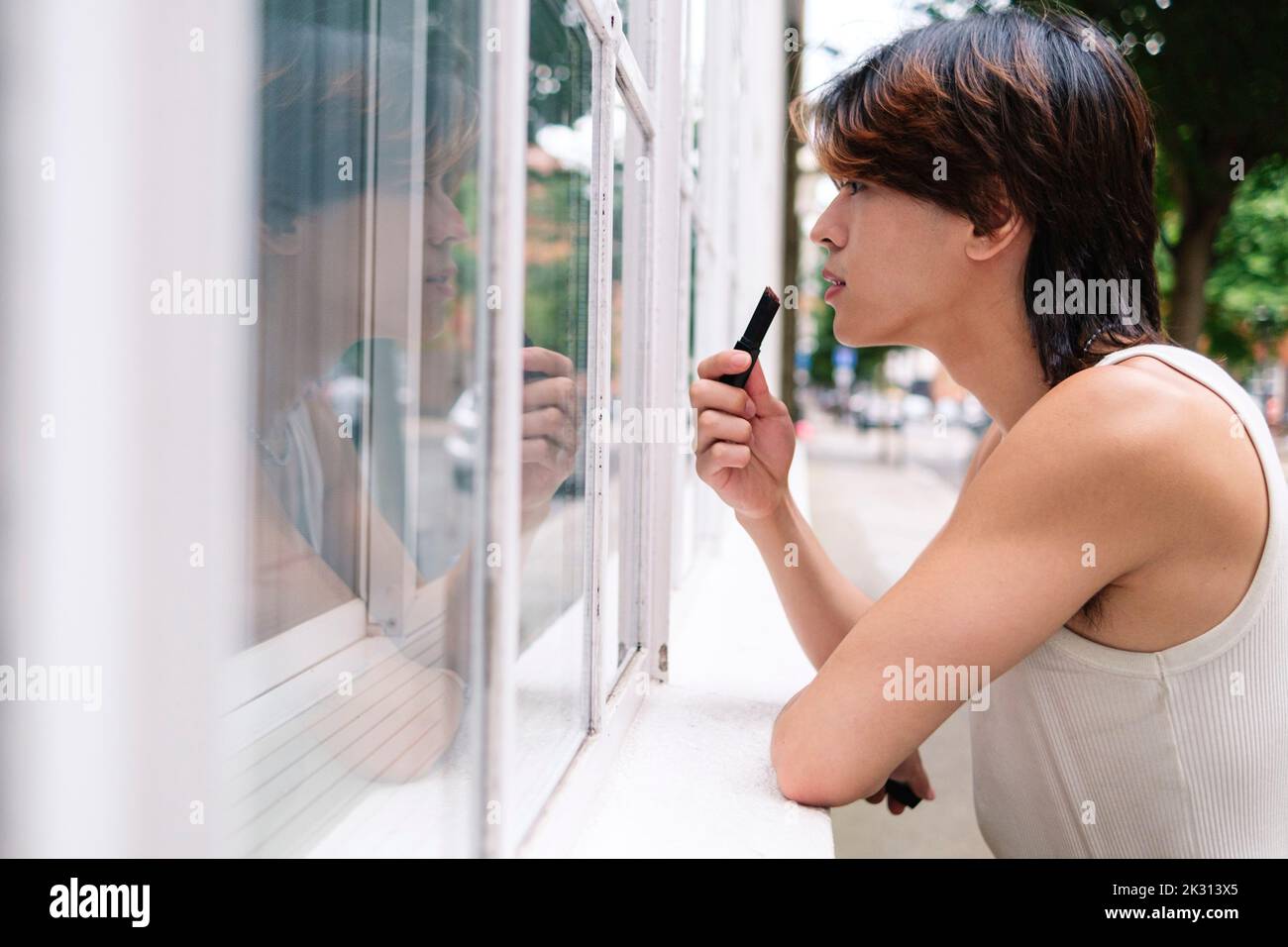 Jeune homme gay appliquant le rouge à lèvres regardant la fenêtre Banque D'Images