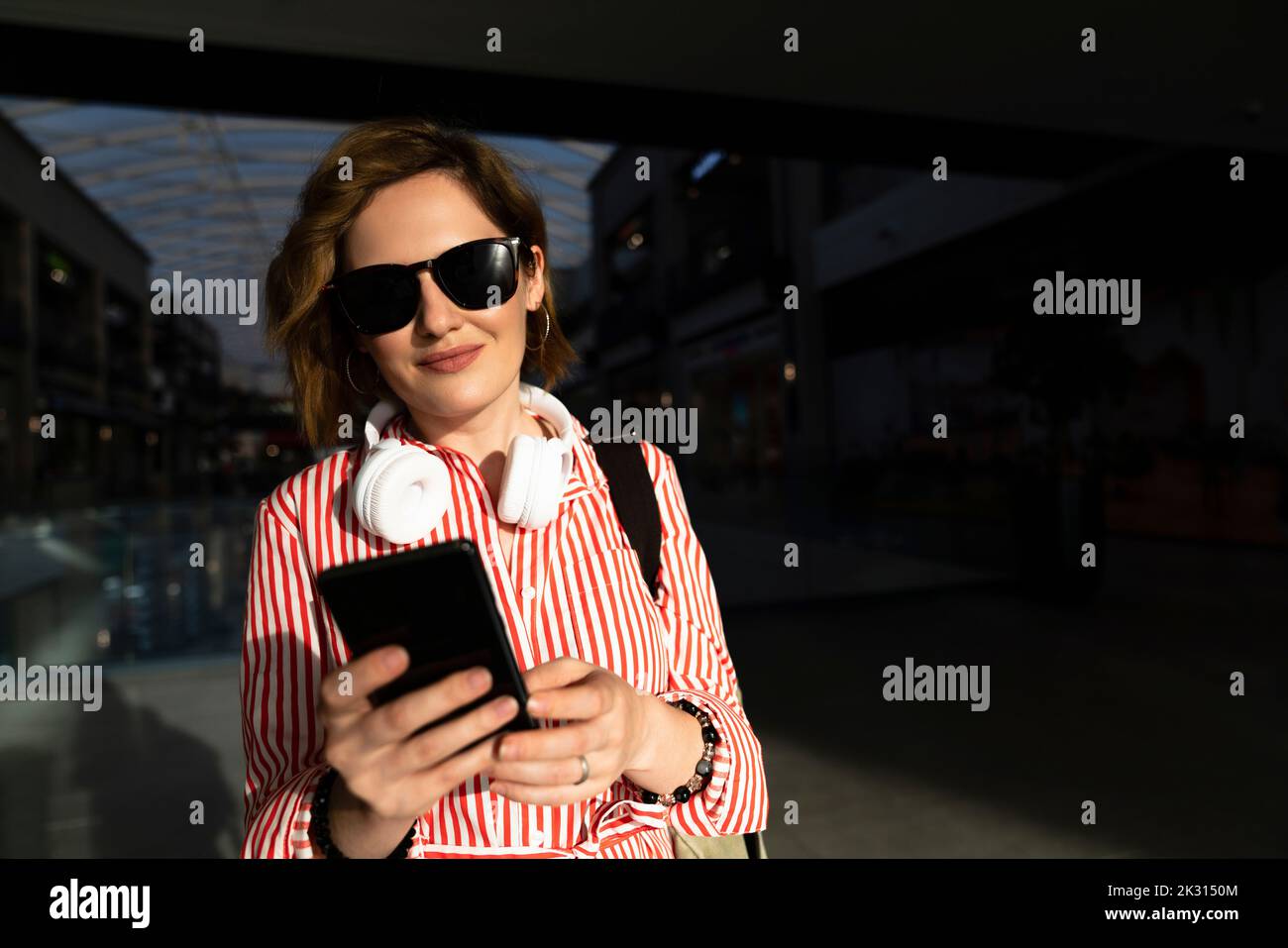 Femme souriante portant des lunettes de soleil à l'aide d'un smartphone dans le centre commercial Banque D'Images